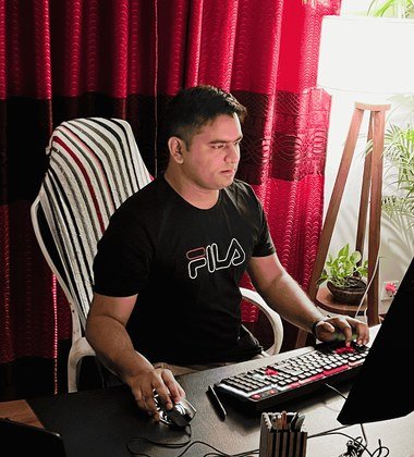 A man working alone at a desk in a home office, one hand on a keyboard and one on a mouse, a red curtain and a floor lamp behind him.